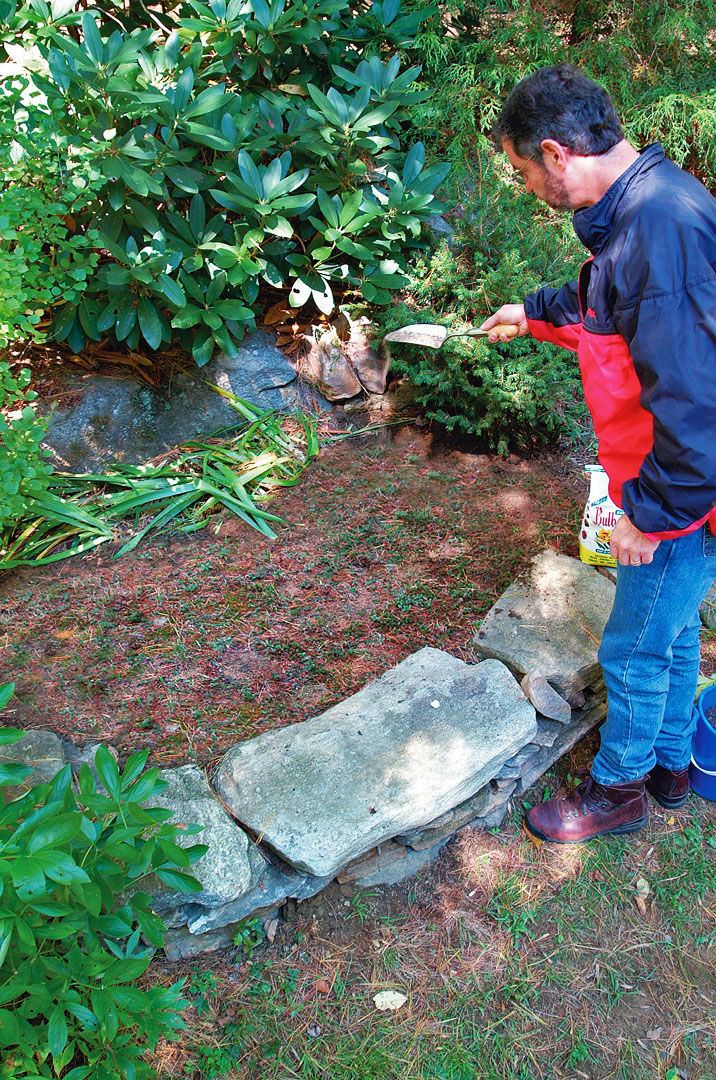 man standing over the rock bordered bulb area holding out a small trowel