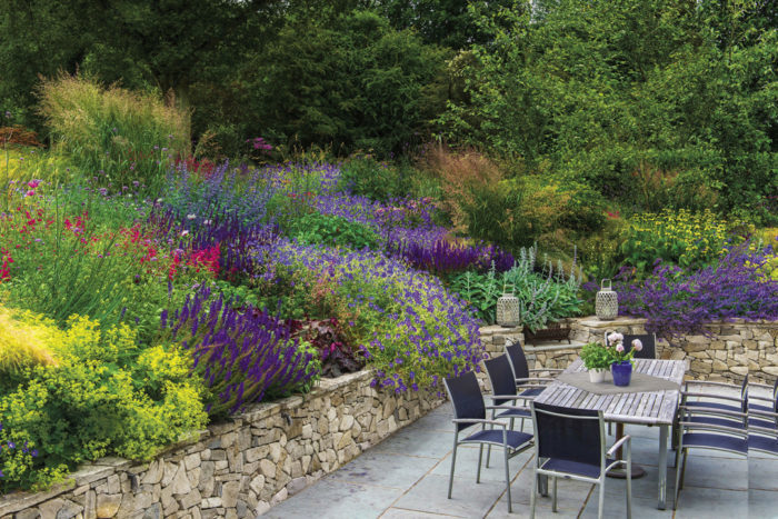 Mounding plants tumbling over a retaining wall’s edge combine with bright spikes of perennial flowers and architectural grasses to wrap a patio in color and texture.