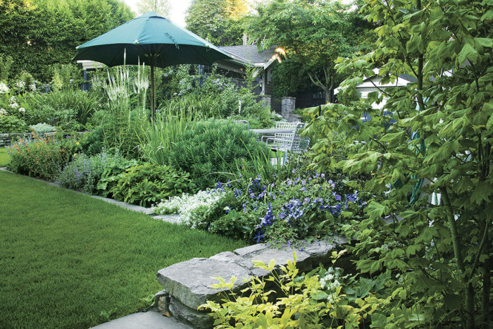 A mature-looking garden full of plants with an umbrella, stone wall and small stone patio