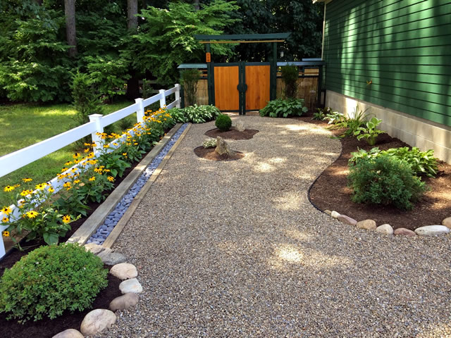 view heading toward the gate with rock path bordered by a fence, flowers and plantings in mulch