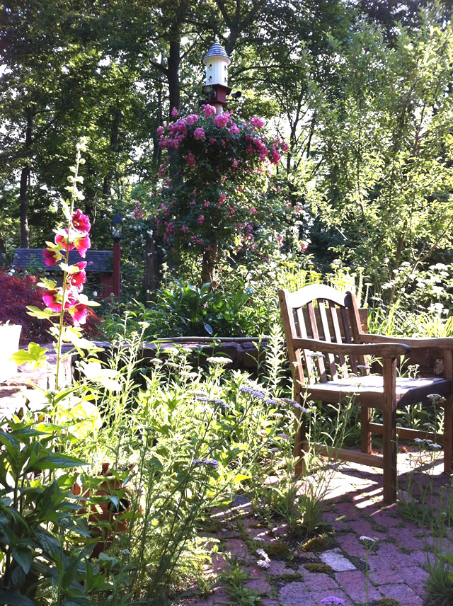 chair on brick, birdhouse on a post with pink roses climbing it, other flowers nearby