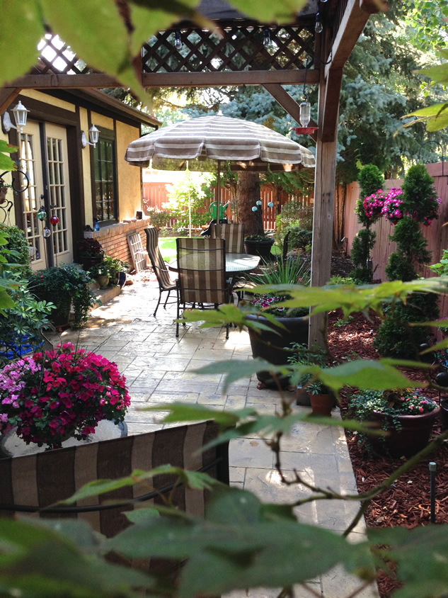 patio longer view with table and chairs, umbrella and trees, shrubs, and large-scale perennials.