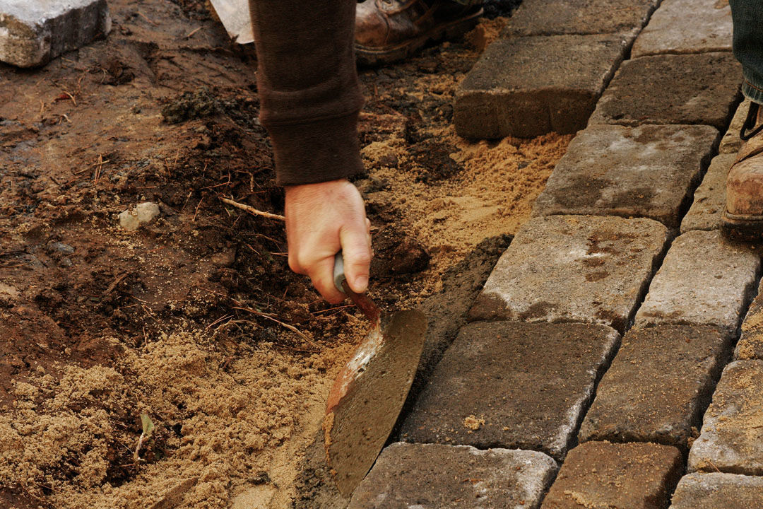 hand using a trowel to scoop some concrete and pack it on an angle along the outside edge of the pavers,