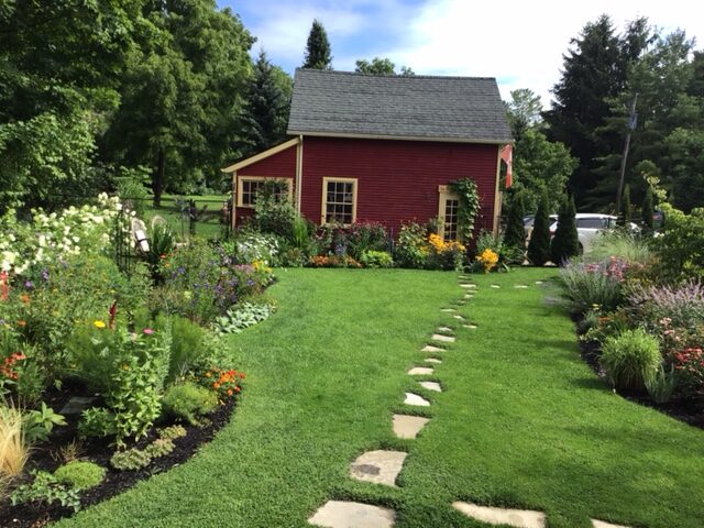 A path through a lawn to a red barn, flanked by flower gardens