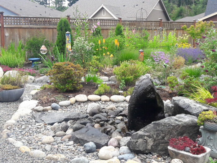 A dark boulder with water tricking over it, with a flower garden behind it.