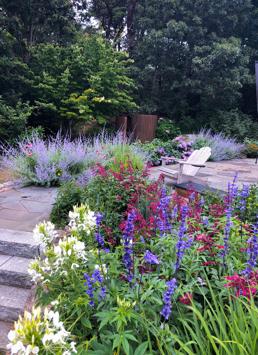 A stone patio with a chair surrounded by purple, blue, burgundy and white flowers