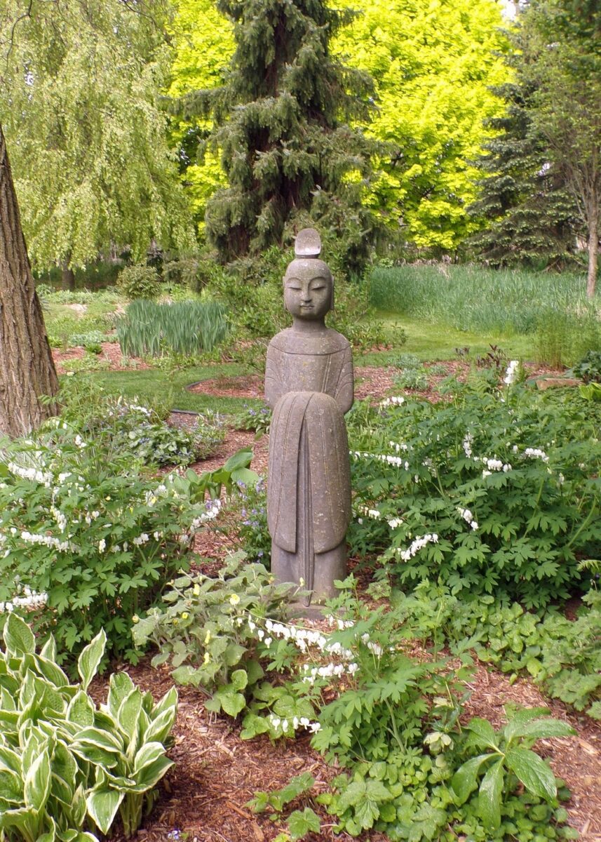 stone statue in the middle of shade plants in bloom
