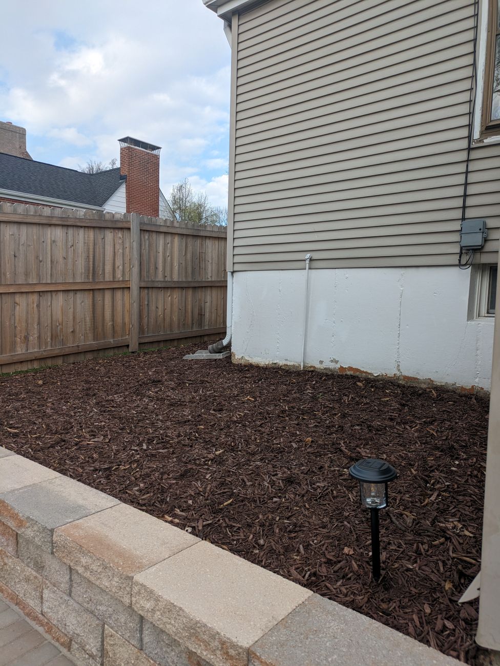 Empty mulched planting bed beside a newly built stone patio and brick house.