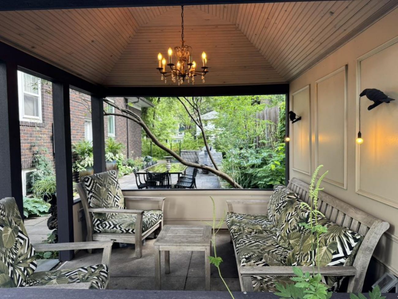 Screened gazebo interior with a vaulted wood ceiling, chandelier, cushioned seating, and garden views.