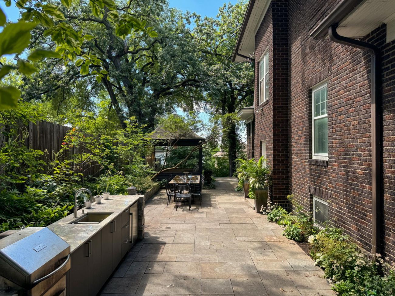 Long bluestone patio with an outdoor kitchen, dining table, layered shade beds, and a screened gazebo.