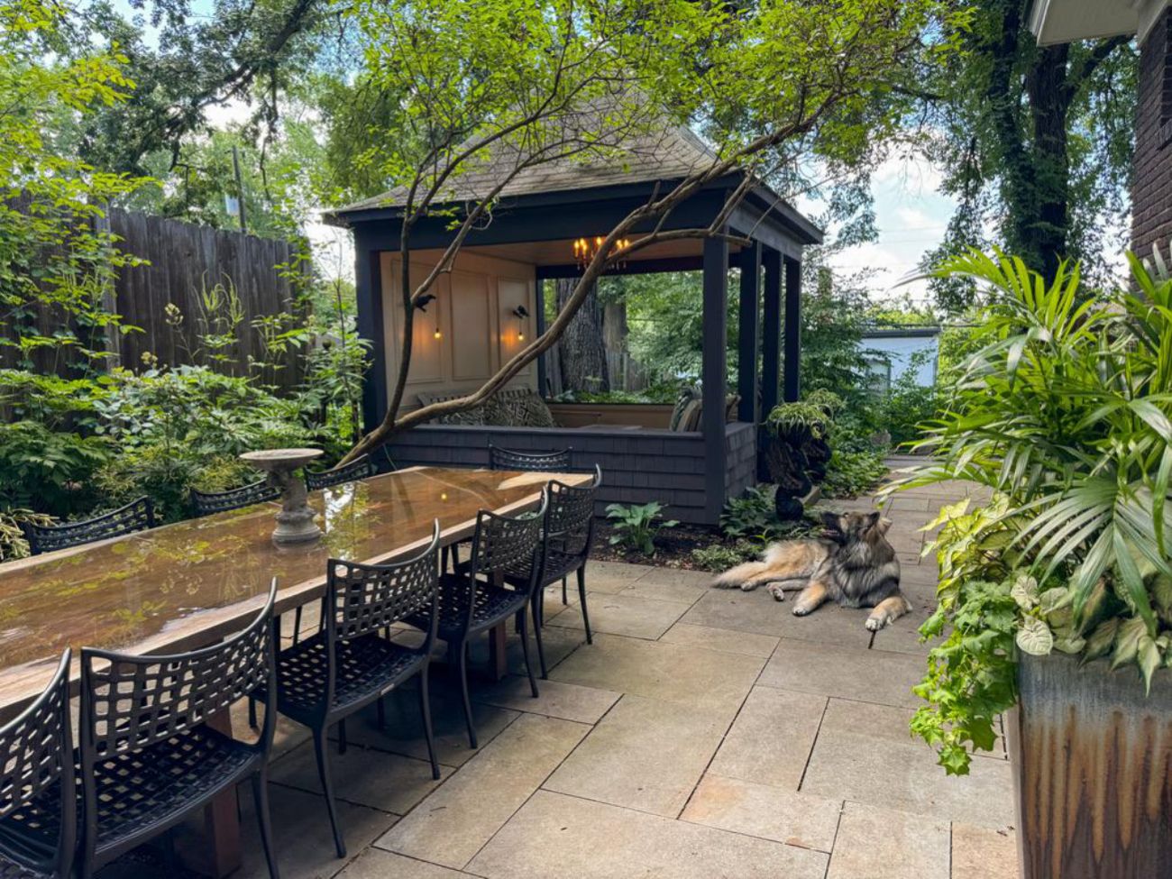 Long outdoor dining table beside a dark screened gazebo surrounded by lush shade plantings.