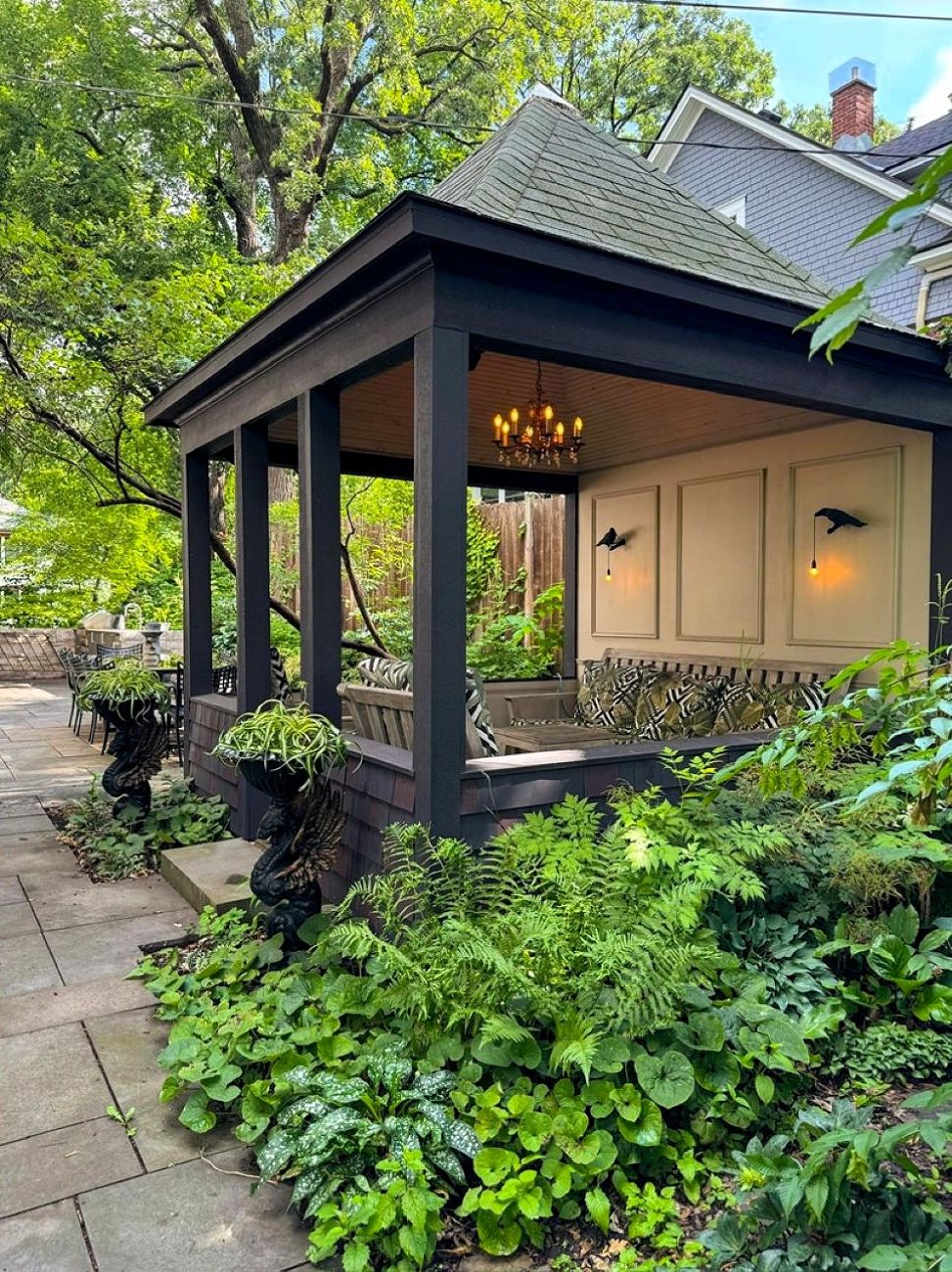 Dark screened gazebo surrounded by layered hostas, ferns, and other shade plants beside a stone patio.